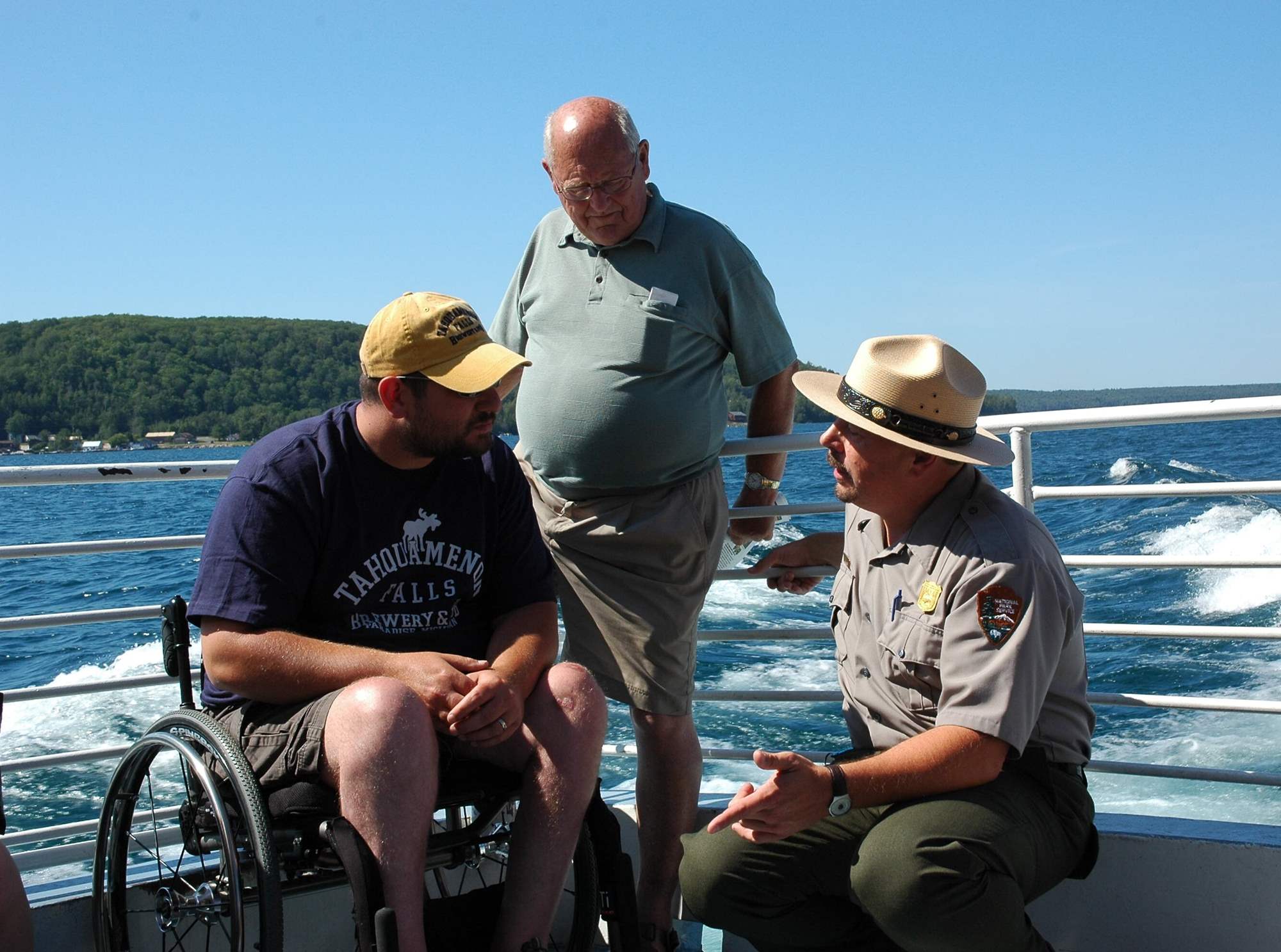 Man in NPS uniform with a shield-shaped badge and broad rim hat kneels on the deck of a boat talking to a man in wheelchair and a standing man.